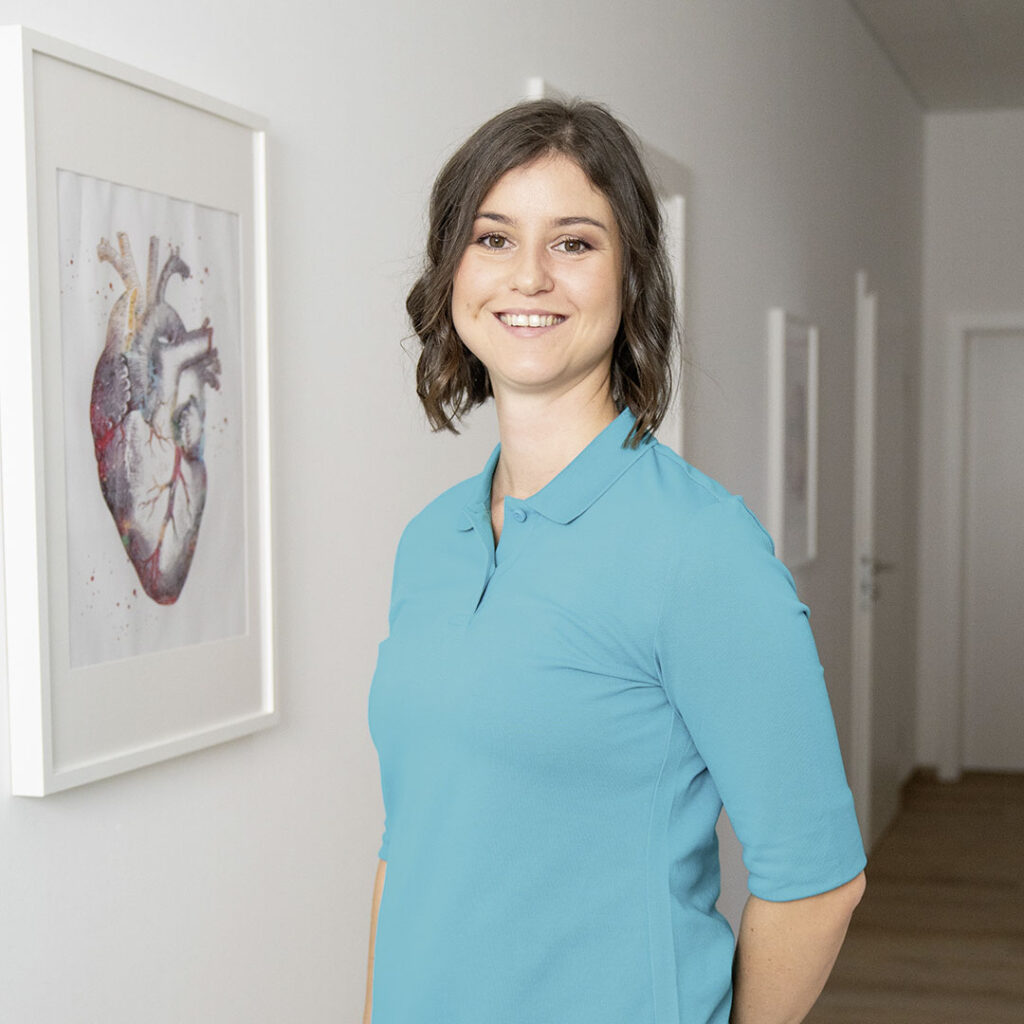 Ina Mayer vor einem Behandlungsraum mit einem Bild, welches das menschliche Herz im Detail zeigt, im Hintergrund.