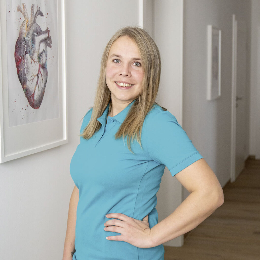 Julia Stockinger vor einem Behandlungsraum mit einem Bild, welches das menschliche Herz im Detail zeigt, im Hintergrund.